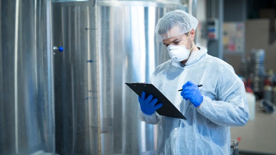 Un homme en blouse blanche et gants bleus tient un clipboard, représentant un fabricant dans un environnement professionnel.
